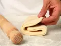 Mani che piegano un impasto per pasta fatta in casa, con un mattarello di legno accanto, cosparso di farina.
