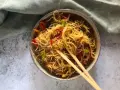 Un piatto invitante di spaghetti di riso con verdure colorate, come peperoni rossi e zucchine, guarnito con basilico fresco e servito con bacchette.