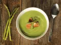 Una ciotola di cremosa crema di asparagi, guarnita con crostini e germogli freschi, accanto a un mazzo di asparagi su un tavolo di legno.