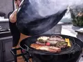 Un uomo solleva il coperchio di una grigliata di carne fumante, rivelando salsicce, pancetta, hamburger e verdure.