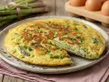 Deliziosa frittata di asparagi selvatici, appena sfornata e guarnita con prezzemolo fresco. Sullo sfondo, un mazzo di asparagi e uova.