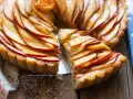 Una deliziosa crostata di mele, con fette di mela lucide disposte a spirale su una base dorata. Una fetta è stata tagliata, rivelando la morbidezza del ripieno.