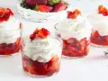 Coppette di fragole con panna montata e pezzetti di fragola fresca in cima. Un cestino di fragole sullo sfondo.