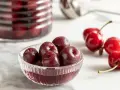 Ciliegie sotto spirito in una ciotola di vetro, con altre ciliegie fresche e un barattolo sullo sfondo.