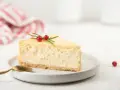 Una fetta di cheesecake yogurt greco, guarnita con bacche rosse e rosmarino, su un piatto bianco con un cucchiaio dorato.
