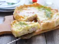 A slice of prosciutto and cheese tart is lifted from the whole tart on a wooden board, garnished with dill.