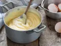 Crema pasticcera veloce in pentola, con cucchiaio di legno che mescola. Guscio d'uovo accanto, pronto per un dolce.