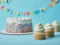 Torta bianca con codette colorate e cupcake decorati con soffice crema al burro, su sfondo azzurro con festoni.