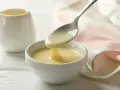 Un cucchiaio versa un denso latte condensato bimby in una ciotola bianca. Sullo sfondo, una piccola brocca e un tovagliolo rosa.