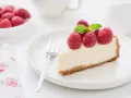 Una fetta di torta fredda allo yogurt guarnita con lamponi freschi e menta, su un piatto bianco con una forchetta.