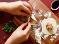 Mani che preparano il ripieno per ravioli su un tagliere di legno, con erba cipollina e salsa di soia a lato.