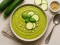 Vellutata di zucchine cremosa, guarnita con basilico fresco, fette di zucchina e pinoli. Accanto, zucchine fresche e un cucchiaio.