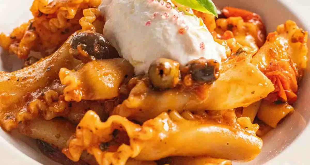Pasta con melanzane, condita con un sugo ricco, olive e una generosa quenelle di ricotta fresca, guarnita con basilico.
