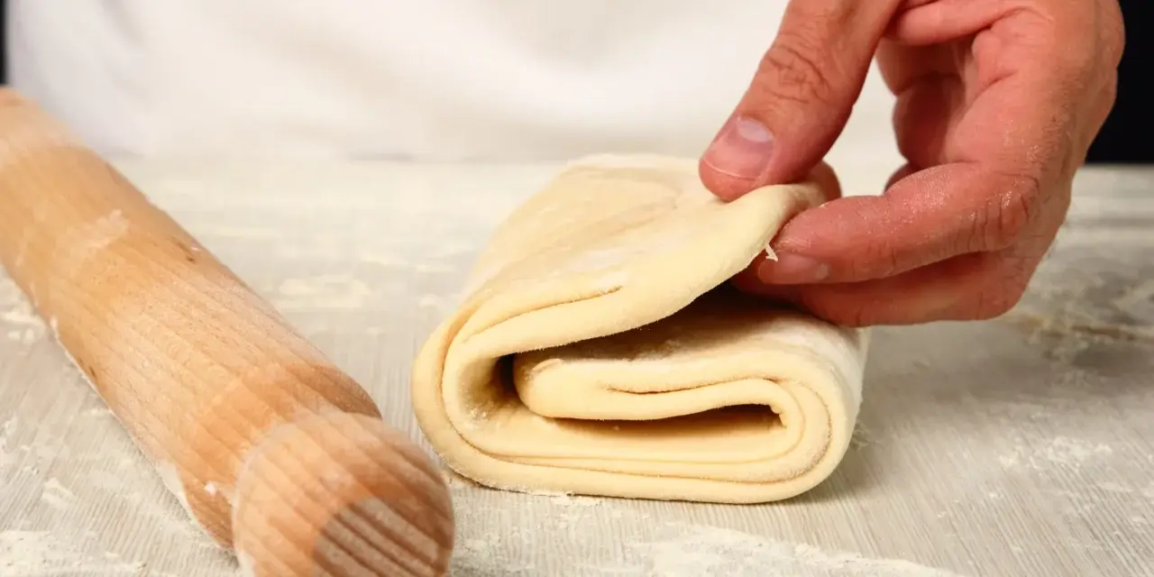 Mani che piegano un impasto per pasta fatta in casa, con un mattarello di legno accanto, cosparso di farina.