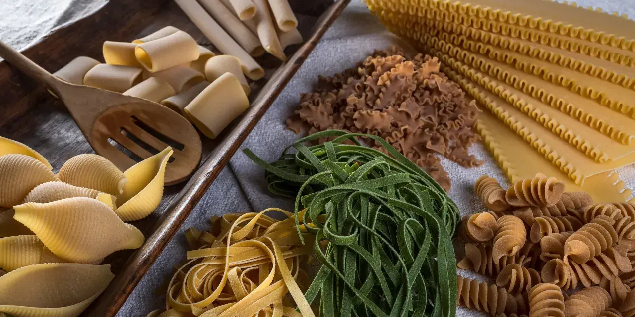 Varietà di pasta cruda, tra cui conchiglioni, paccheri, fettuccine verdi e gialle, lasagne e fusilli, pronte per essere condite con i migliori sughi per pasta.