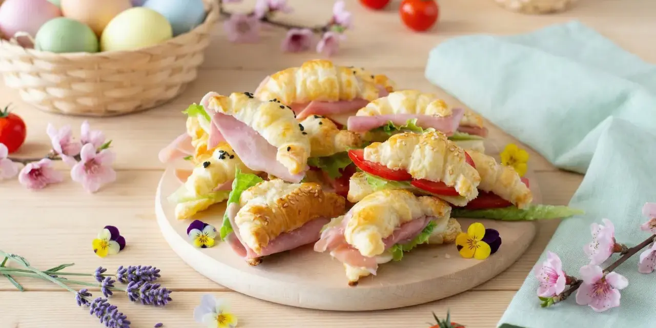 Delizioso antipasto di Pasqua con croissant farciti, uova colorate in un cesto e fiori primaverili.