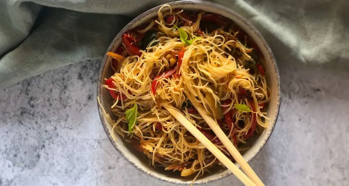 Un piatto invitante di spaghetti di riso con verdure colorate, come peperoni rossi e zucchine, guarnito con basilico fresco e servito con bacchette.
