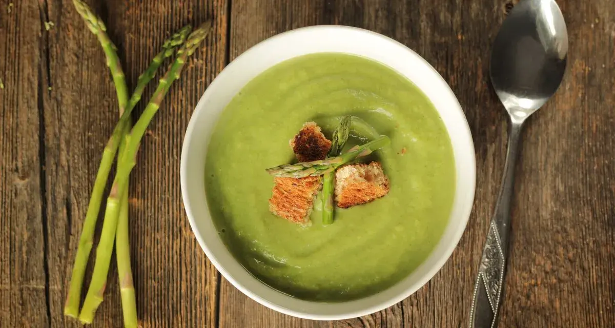 Una ciotola di cremosa crema di asparagi, guarnita con crostini e germogli freschi, accanto a un mazzo di asparagi su un tavolo di legno.