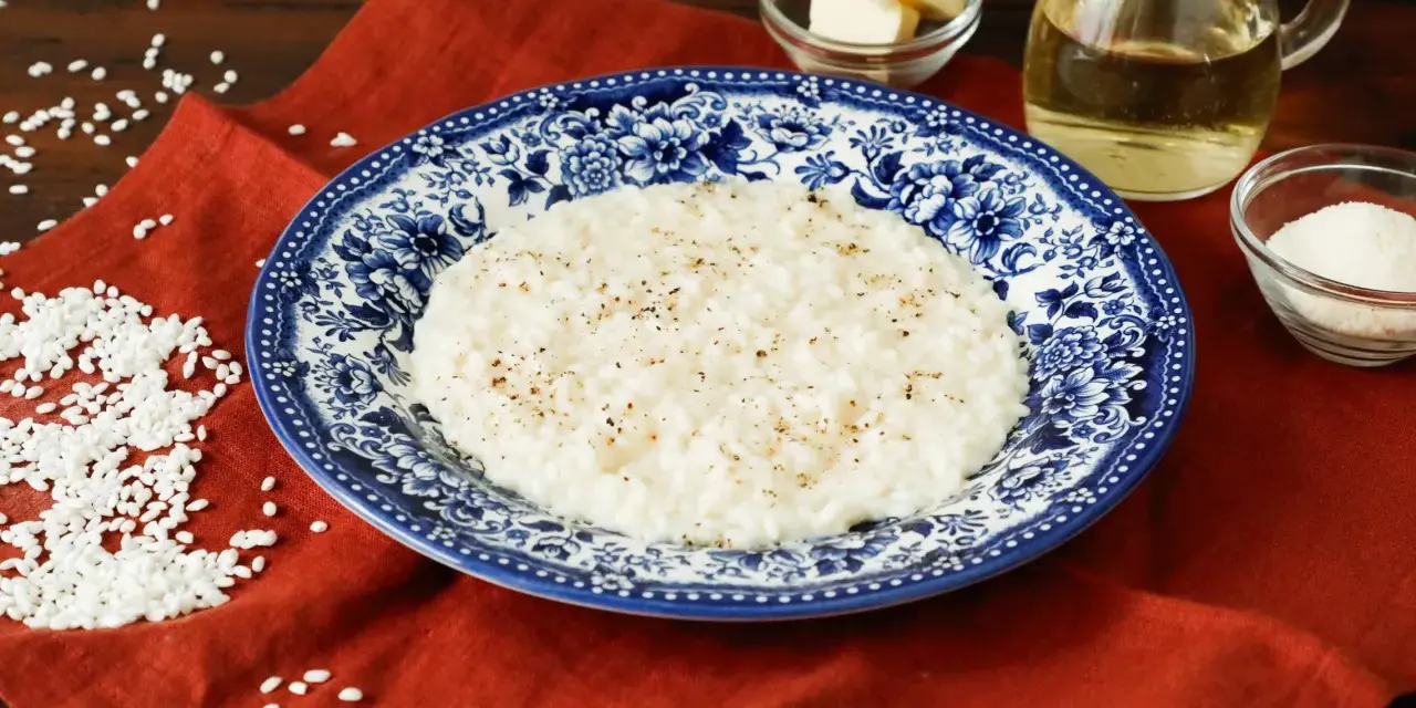 Un cremoso riso in bianco, condito con pepe nero, servito su un piatto decorato. Accanto, burro, olio e parmigiano grattugiato, ingredienti per un piatto semplice ma gustoso.