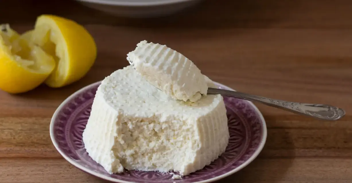 Ricotta fatta in casa su un piatto viola, con limone tagliato a metà sullo sfondo. Un cucchiaino preleva una porzione cremosa.