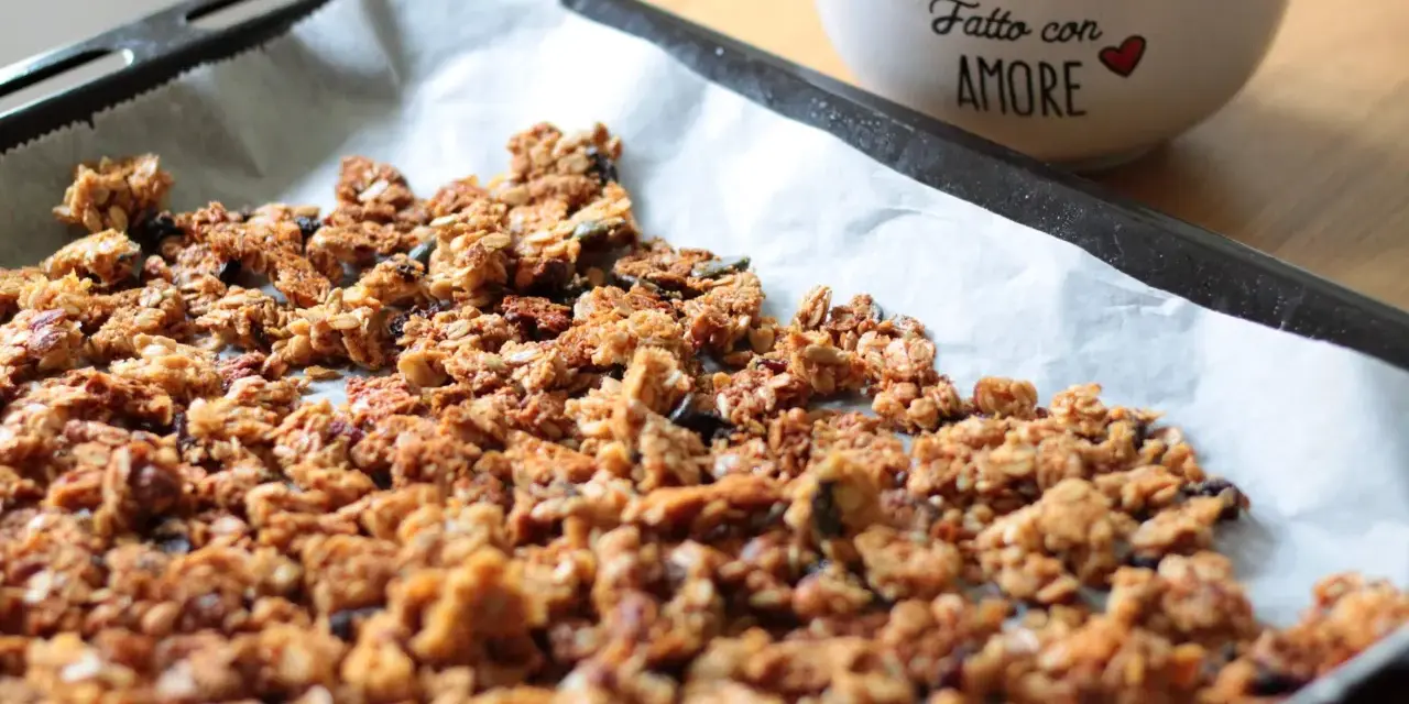 Granola fatta in casa appena sfornata su carta da forno, con una ciotola di frutta secca sullo sfondo.