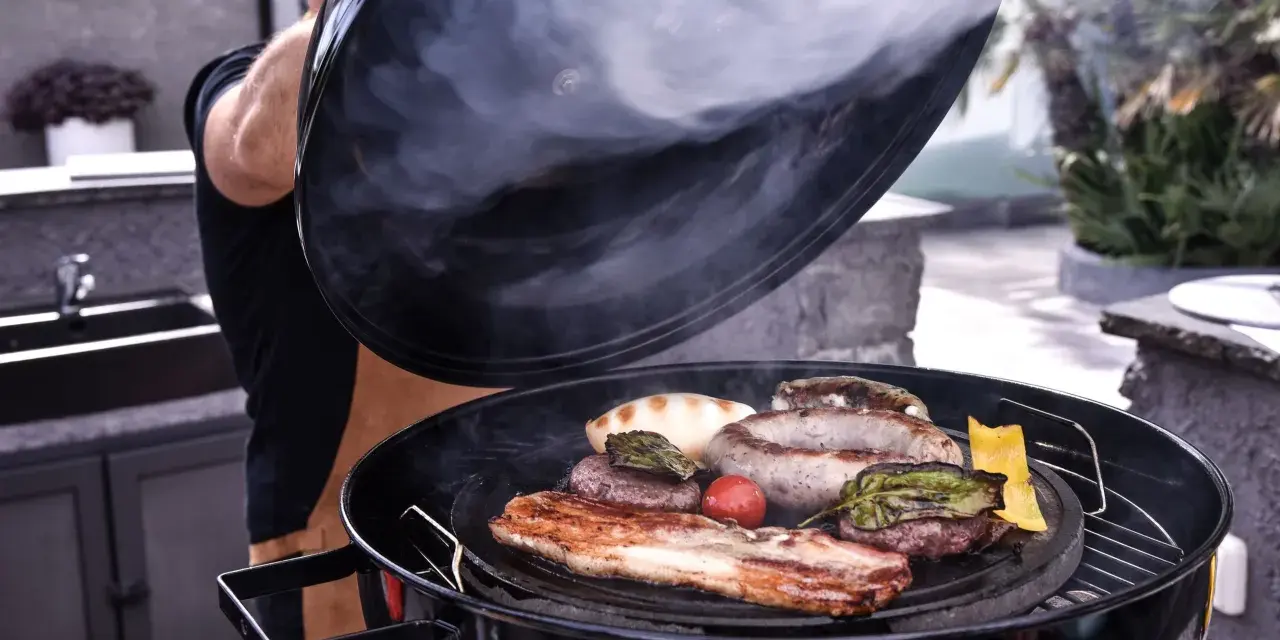 Un uomo solleva il coperchio di una grigliata di carne fumante, rivelando salsicce, pancetta, hamburger e verdure.