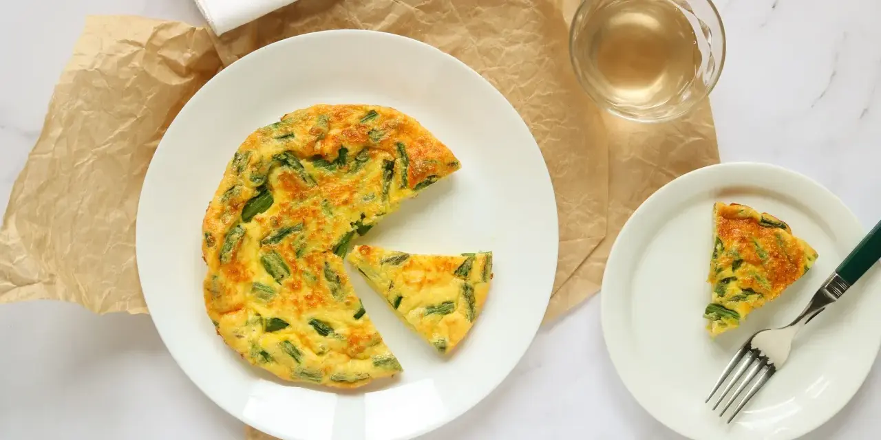 Deliziosa frittata di asparagi al forno, servita su un piatto bianco con una fetta a parte e un bicchiere di vino.