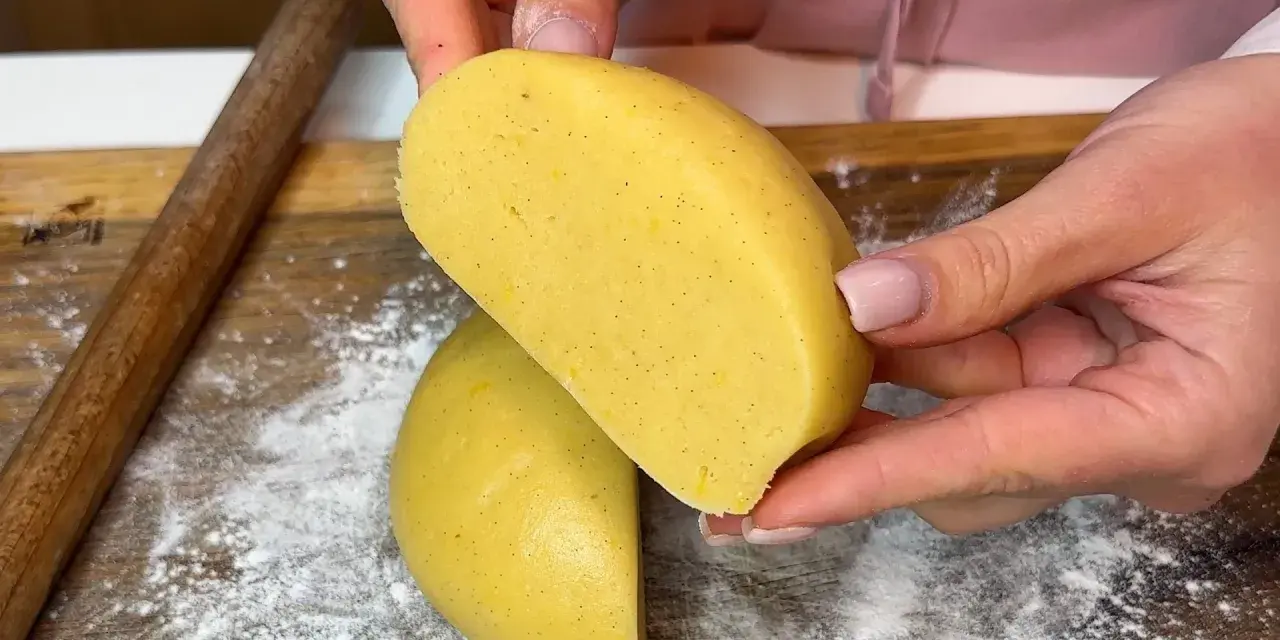 Mani che tengono un impasto giallo, profumato alla vaniglia, pronto per diventare una deliziosa pasta frolla senza burro.
