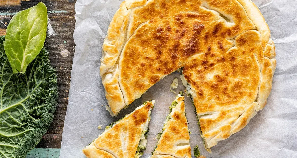 Torta salata con pasta sfoglia dorata, ripiena di verdure. Accanto, foglie di cavolo nero fresco.