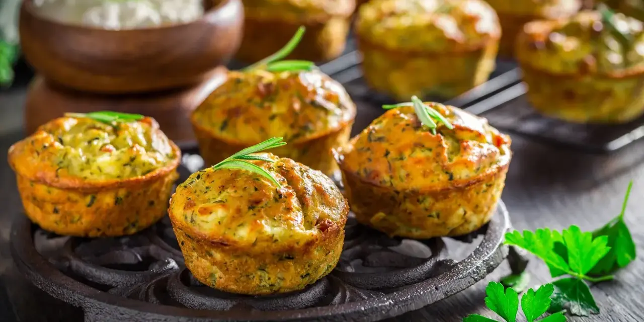 Tortini di zucchine dorati e fragranti, guarniti con rosmarino, su un piatto di ghisa. Sullo sfondo, una ciotola di formaggio fresco.