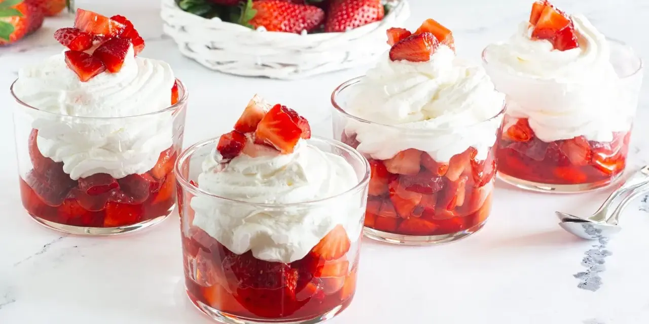 Coppette di fragole con panna montata e pezzetti di fragola fresca in cima. Un cestino di fragole sullo sfondo.