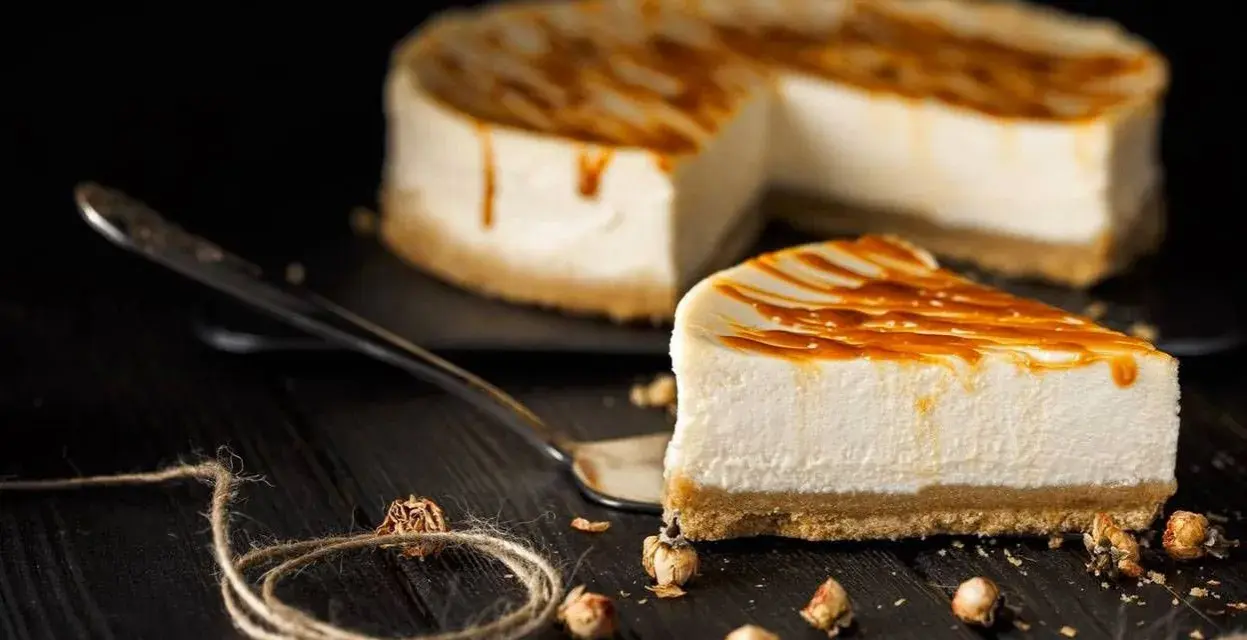 Una fetta di cheesecake senza panna, con glassa caramellata, su un piatto da portata. Sullo sfondo, la torta intera.
