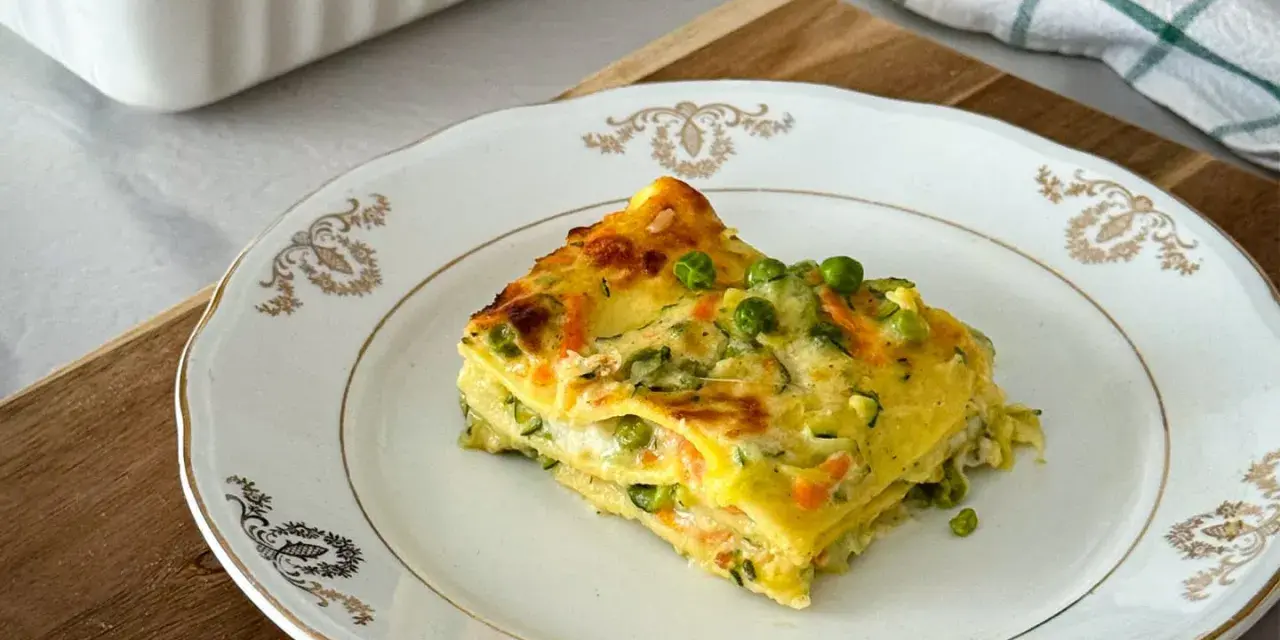 Una fetta di lasagne di verdure, con piselli e carote, su un piatto bianco decorato.