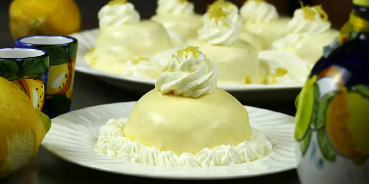 Dolcetti rotondi con glassa al limone e panna montata, guarniti con scorza di limone. Sullo sfondo, limoni freschi e bicchierini decorati.