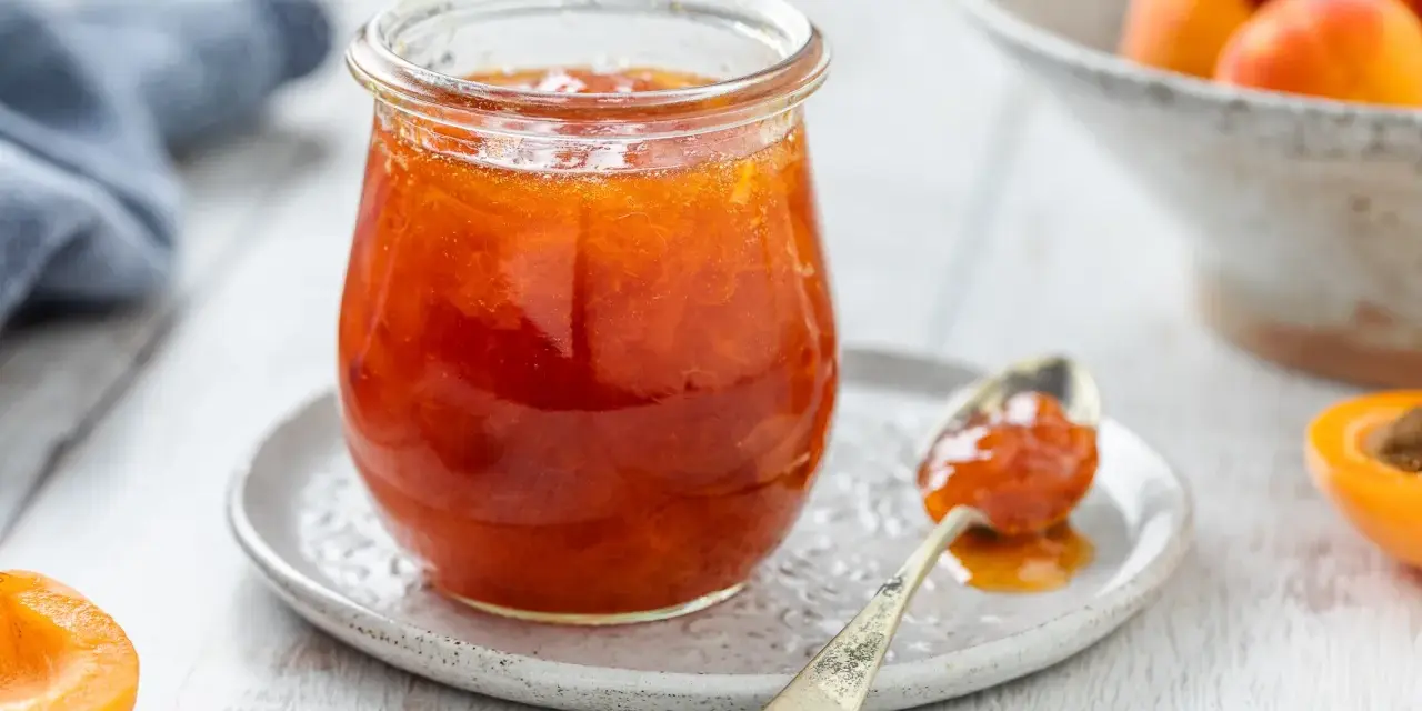 Vasetto di marmellata di albicocche, con un cucchiaino carico e albicocche fresche sullo sfondo.