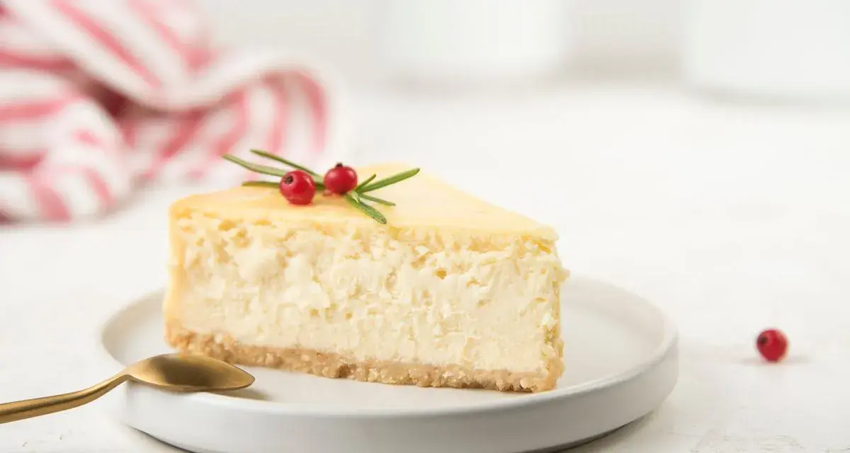 Una fetta di cheesecake yogurt greco, guarnita con bacche rosse e rosmarino, su un piatto bianco con un cucchiaio dorato.