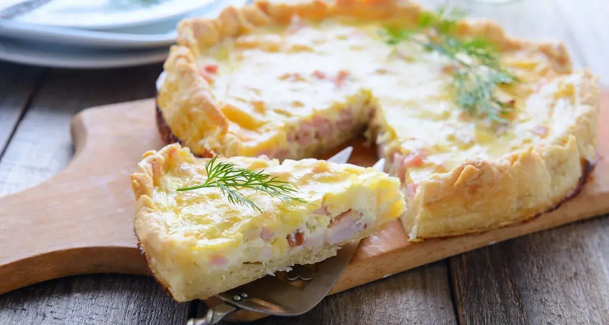 A slice of prosciutto and cheese tart is lifted from the whole tart on a wooden board, garnished with dill.