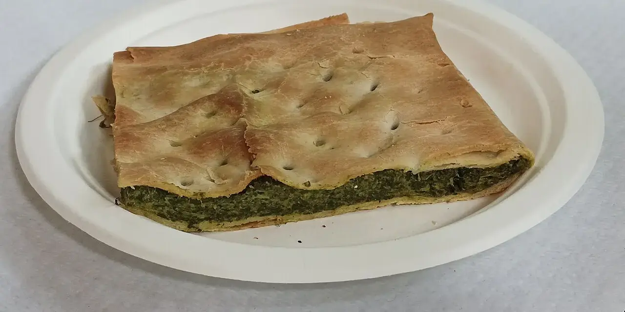 Una fetta di torta pasqualina originale genovese, con pasta sfoglia dorata e ripieno verde di spinaci e ricotta, servita su un piatto bianco.