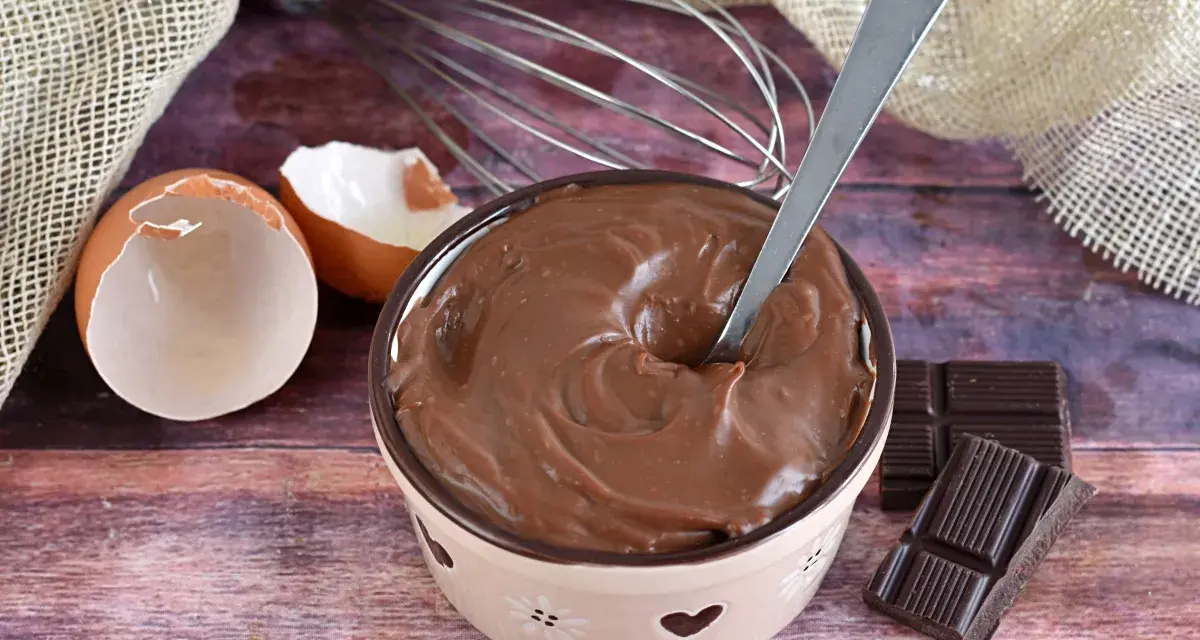Crema pasticcera al cioccolato, liscia e invitante, pronta per essere gustata. Uova, frusta e cioccolato, ingredienti per un dolce momento.