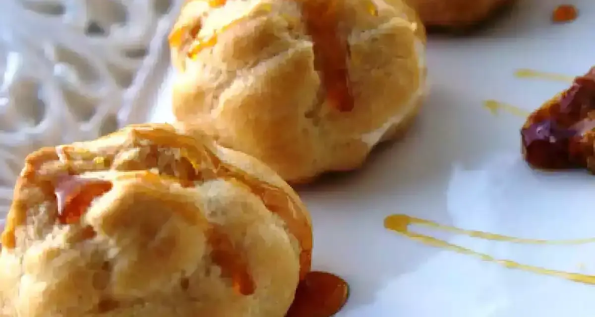 Bignè alla crema soffici e dorati, bagnati da un filo di caramello lucido su un piatto bianco.