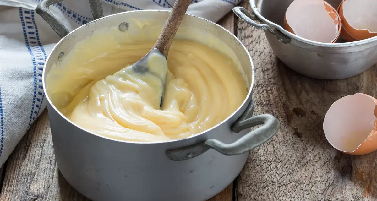 Crema pasticcera veloce in pentola, con cucchiaio di legno che mescola. Guscio d'uovo accanto, pronto per un dolce.