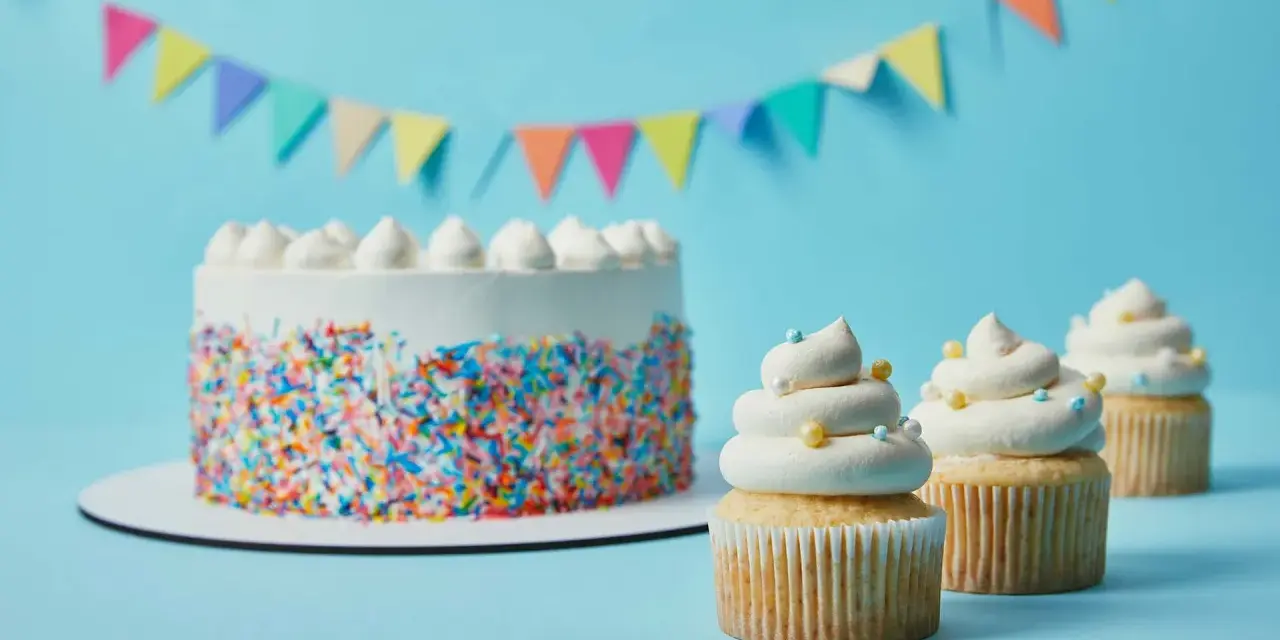 Torta bianca con codette colorate e cupcake decorati con soffice crema al burro, su sfondo azzurro con festoni.