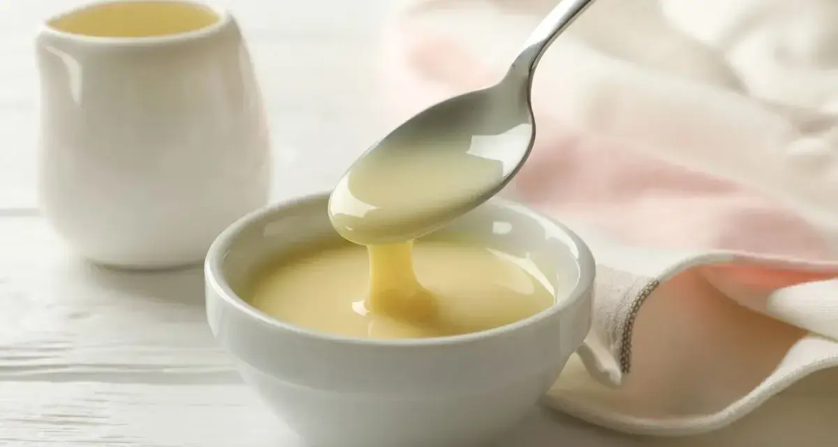 Un cucchiaio versa un denso latte condensato bimby in una ciotola bianca. Sullo sfondo, una piccola brocca e un tovagliolo rosa.