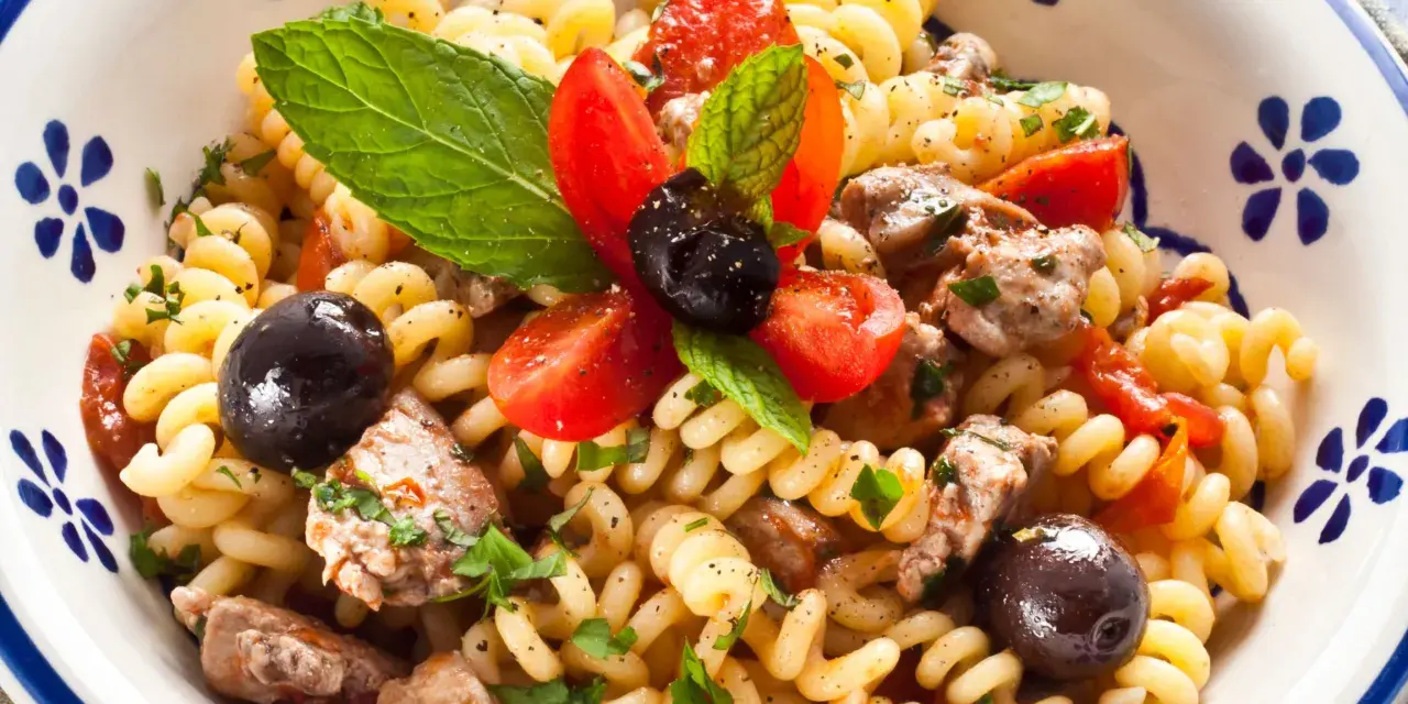 Pasta con pesce spada, pomodorini, olive nere e menta fresca in una ciotola decorata. Un piatto estivo che profuma di mare.