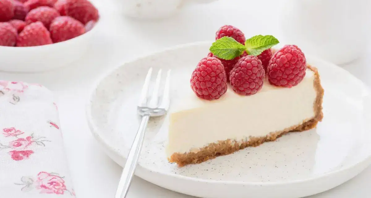 Una fetta di torta fredda allo yogurt guarnita con lamponi freschi e menta, su un piatto bianco con una forchetta.