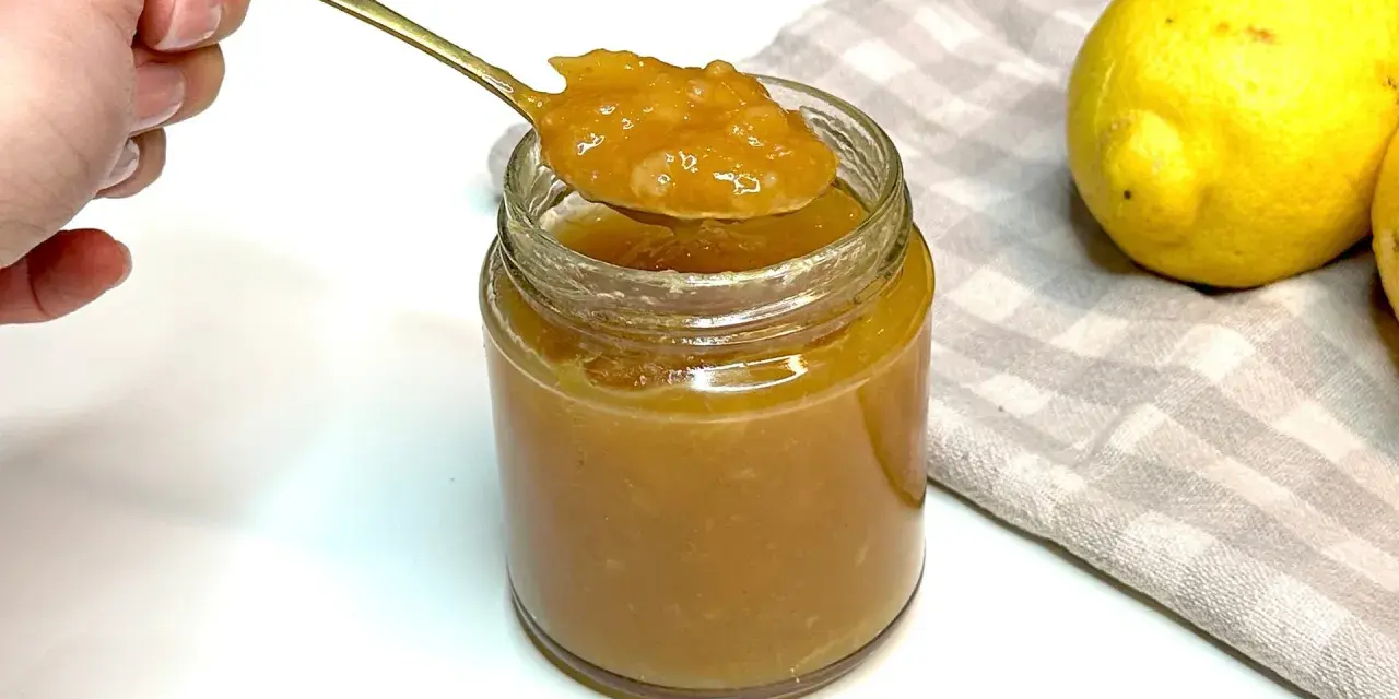 Un cucchiaio d'oro raccoglie la densa marmellata di limoni da un barattolo di vetro. Due limoni freschi riposano su un tovagliolo a quadretti.