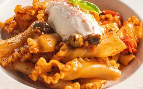 Pasta con melanzane, condita con un sugo ricco, olive e una generosa quenelle di ricotta fresca, guarnita con basilico.
