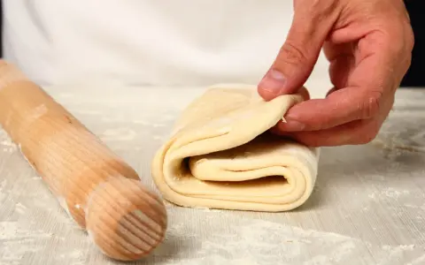 Mani che piegano un impasto per pasta fatta in casa, con un mattarello di legno accanto, cosparso di farina.