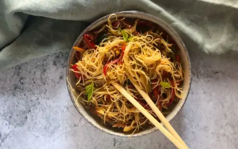 Un piatto invitante di spaghetti di riso con verdure colorate, come peperoni rossi e zucchine, guarnito con basilico fresco e servito con bacchette.