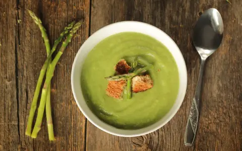 Una ciotola di cremosa crema di asparagi, guarnita con crostini e germogli freschi, accanto a un mazzo di asparagi su un tavolo di legno.